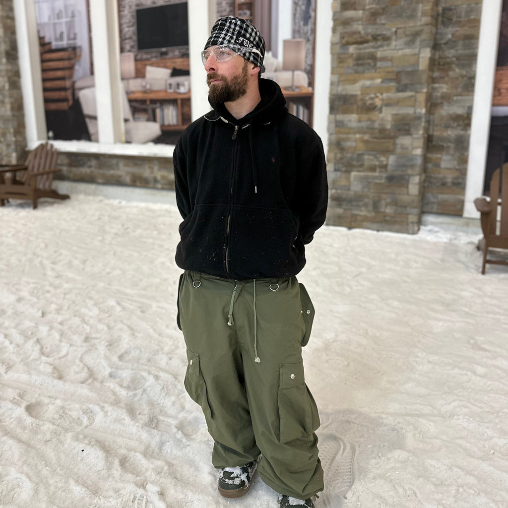 Person wearing a black jacket and green pants standing in a snowy area with a stone building in the background.