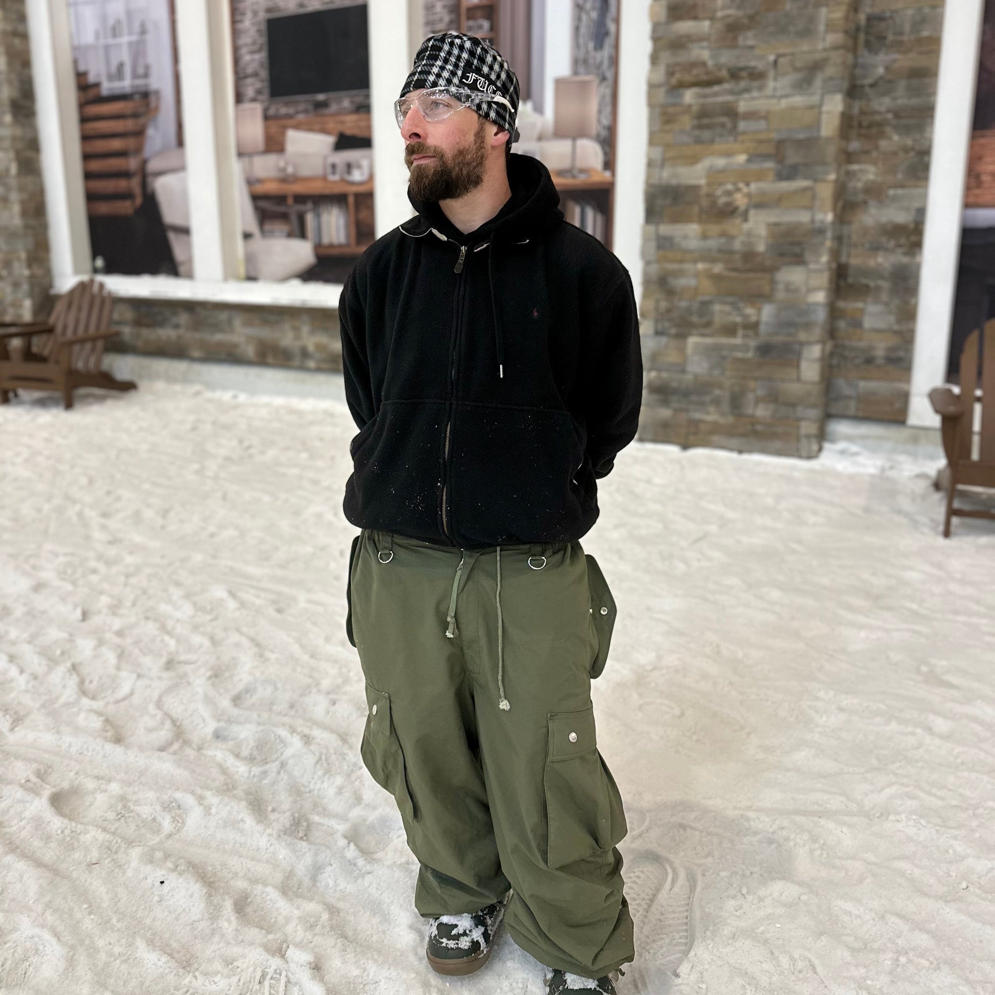 Person wearing a black jacket and green pants standing in a snowy area with a stone building in the background.
