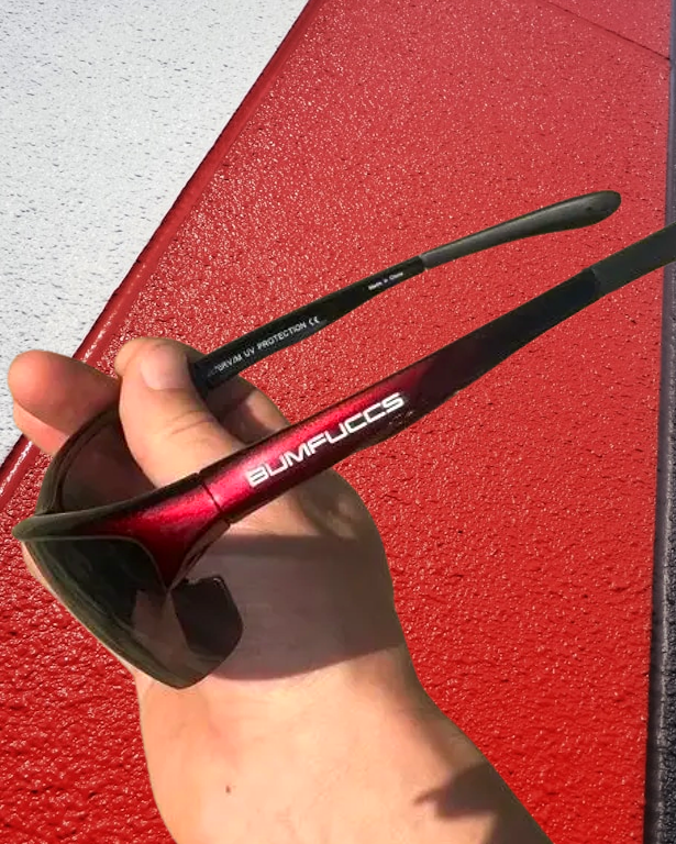 A hand holding a pair of BUMFUCCS Tactical Speed Sunglasses with a red and black frame, featuring UV400 protection lenses, against a red and white checkered background.